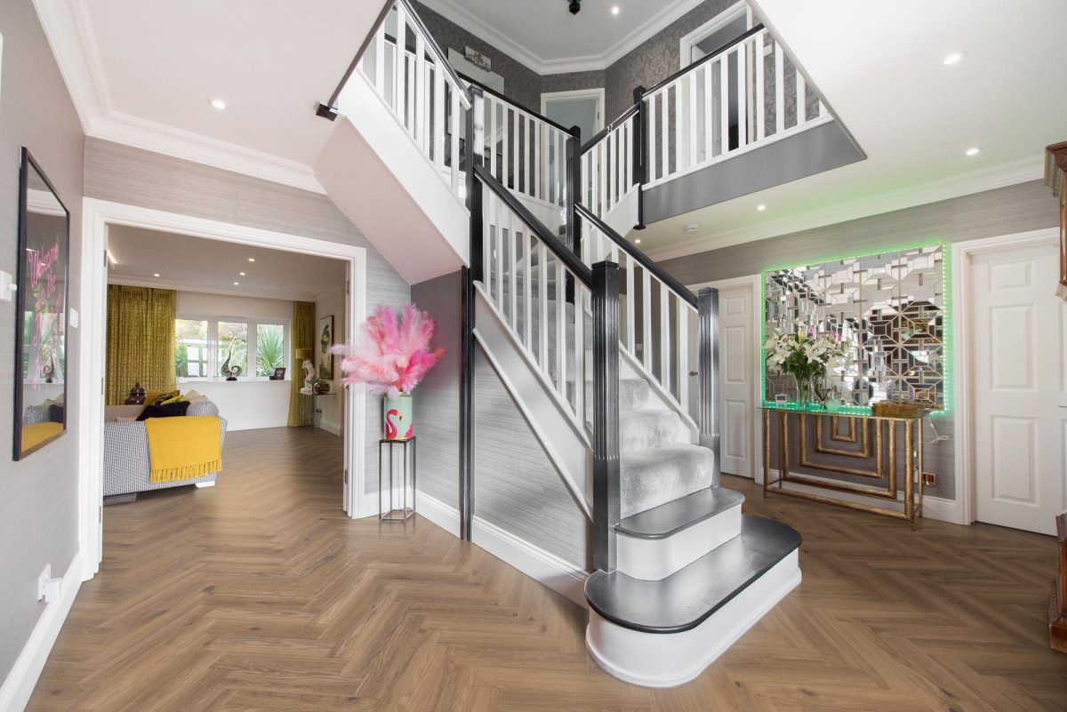 A general interior view of a large grand staircase in a hallway with parquet flooring, vase with pink feathers, gold console table, mirror with LED lighting strip looking through to the lounge